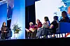 Conference panel with several speakers seated on stage in front of a large projected screen.