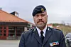 Militæroffiser i mørk uniform med medaljer står foran lave bygninger.