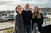 Tre personer står og smiler foran en småbåthavn ved kysten.