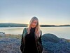 Blonde woman standing on a rocky shoreline with calm water and distant hills at sunset.
