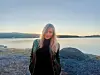 Blonde woman standing on a rocky shoreline with calm water and distant hills at sunset.