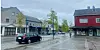 Regnvåt gate med biler, fotgjengere og butikker i et lite sentrum.