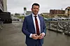 Man in a blue suit stands on a wet harbour walkway with boats and buildings behind him.