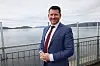Man in a blue suit standing by a waterfront railing with water and hills in the background.