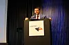 Man speaking at a podium with a Bankers logo on the front at a lit stage event.