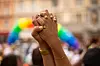 Hender løftet foran en Pride-parade, med regnbueflagg og publikum i bakgrunnen.
