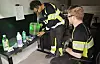 To arbeidere ved en benk med flasker, beholdere og grønn plastbøtte i et innendørs arbeidsrom.