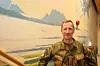 En soldat står foran et veggmaleri med fjell, vann og en isbre.