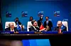Delegates seated at a summit table with microphones, flags and a blue branded backdrop.
