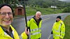 Tre personer i gule refleksjakker står ved en landevei med grønt landskap i bakgrunnen.