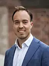 Portrett av Emil Bremnes i blå dressjakke og hvit skjorte foran en uskarp murvegg.