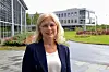 En kvinne står smilende foran et moderne kontorbygg med glass og grøntanlegg.
