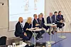 Paneldeltakere sitter ved høye bord foran en projisert presentasjonsskjerm.