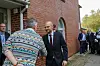 To menn møtes utenfor en rød murbygning, med en gruppe personer i bakgrunnen.