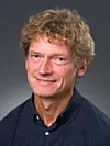 Portrett av en smilende mann med krøllete hår og mørk skjorte.