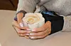 Hender holder en kopp cappuccino med melkeskum på et trebord.