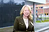 Smilende kvinne i mørk jakke foran moderne bygninger og en gangvei.