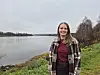 Smilende kvinne i rutete skjorte ved en innsjø under overskyet himmel.