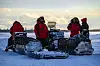 Kanadiske rangere i røde vinterklær samles ved snøscootere i et snødekt arktisk landskap.