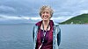 Gunhild Hoogensen Gjørv smiler foran en fjord med grønne åser og overskyet himmel.